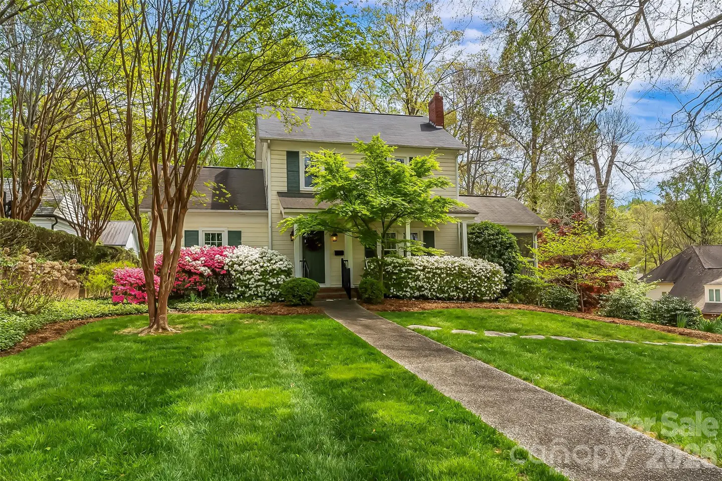 Two-story beige house with green shutters, surrounded by lush green lawn, trees, and colorful flowering bushes. A stone walkway leads to the front door.