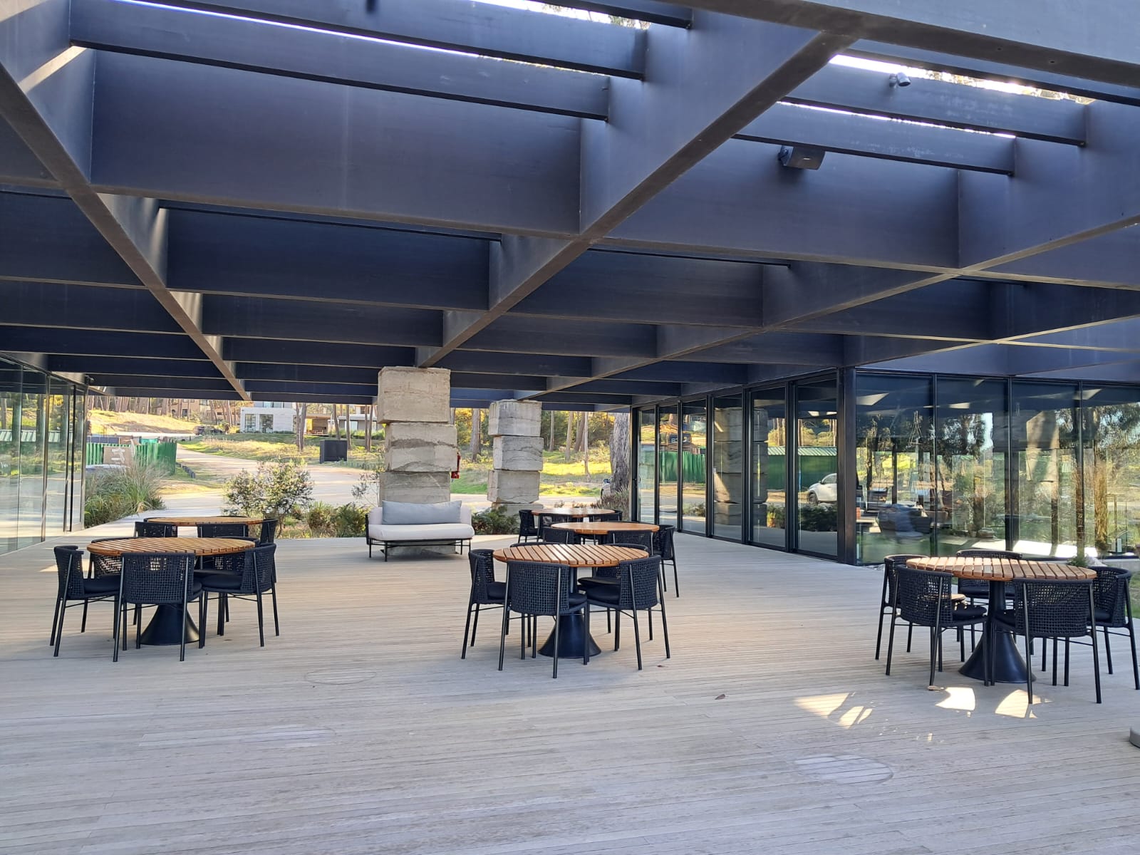 Outdoor patio with round tables, black chairs, and a gray sofa under a modern black pergola. Large windows line the back, overlooking a green landscape.