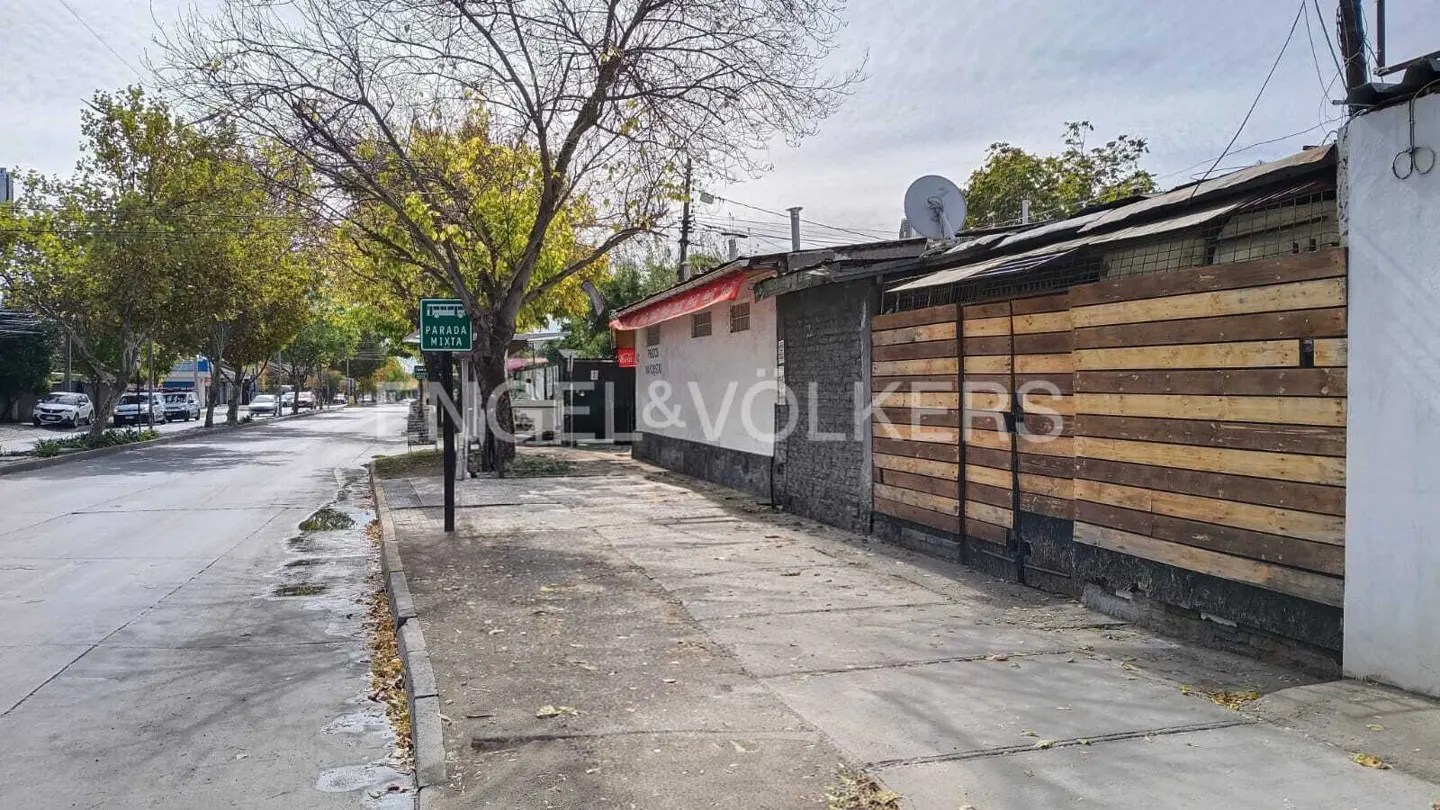 Street view with a sidewalk, trees, and buildings. A wooden fence is on the right. A street sign is on the left.
