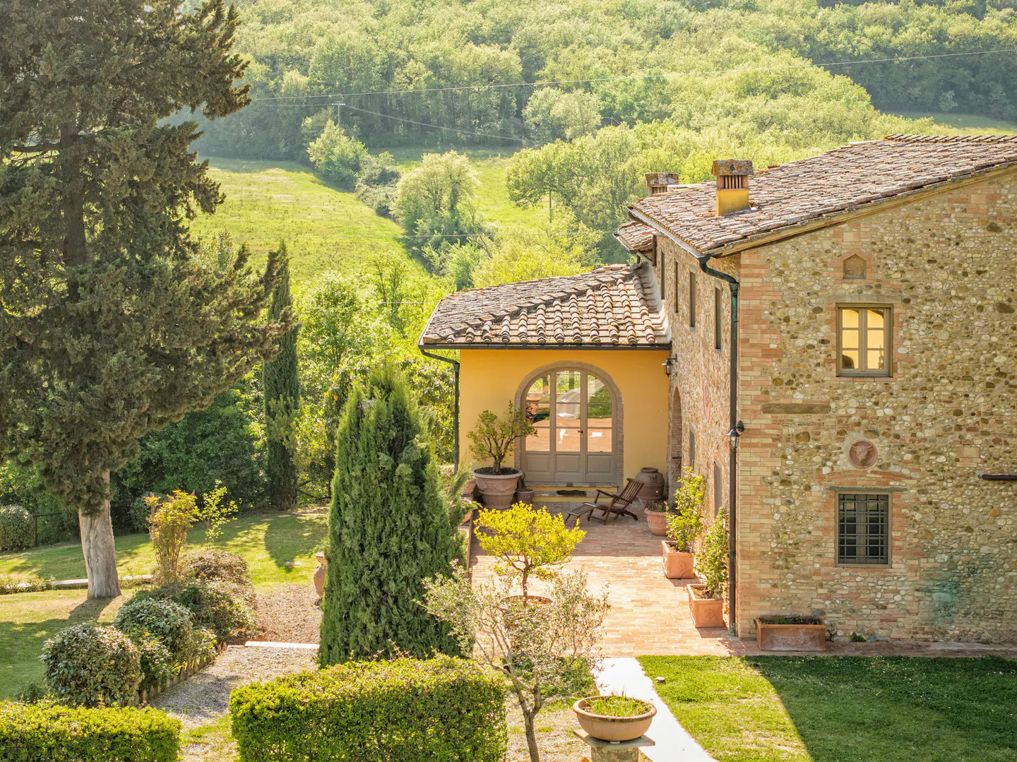 Exterior of a stone house with a tile roof, yellow arched doorway, and green trees in the background.
