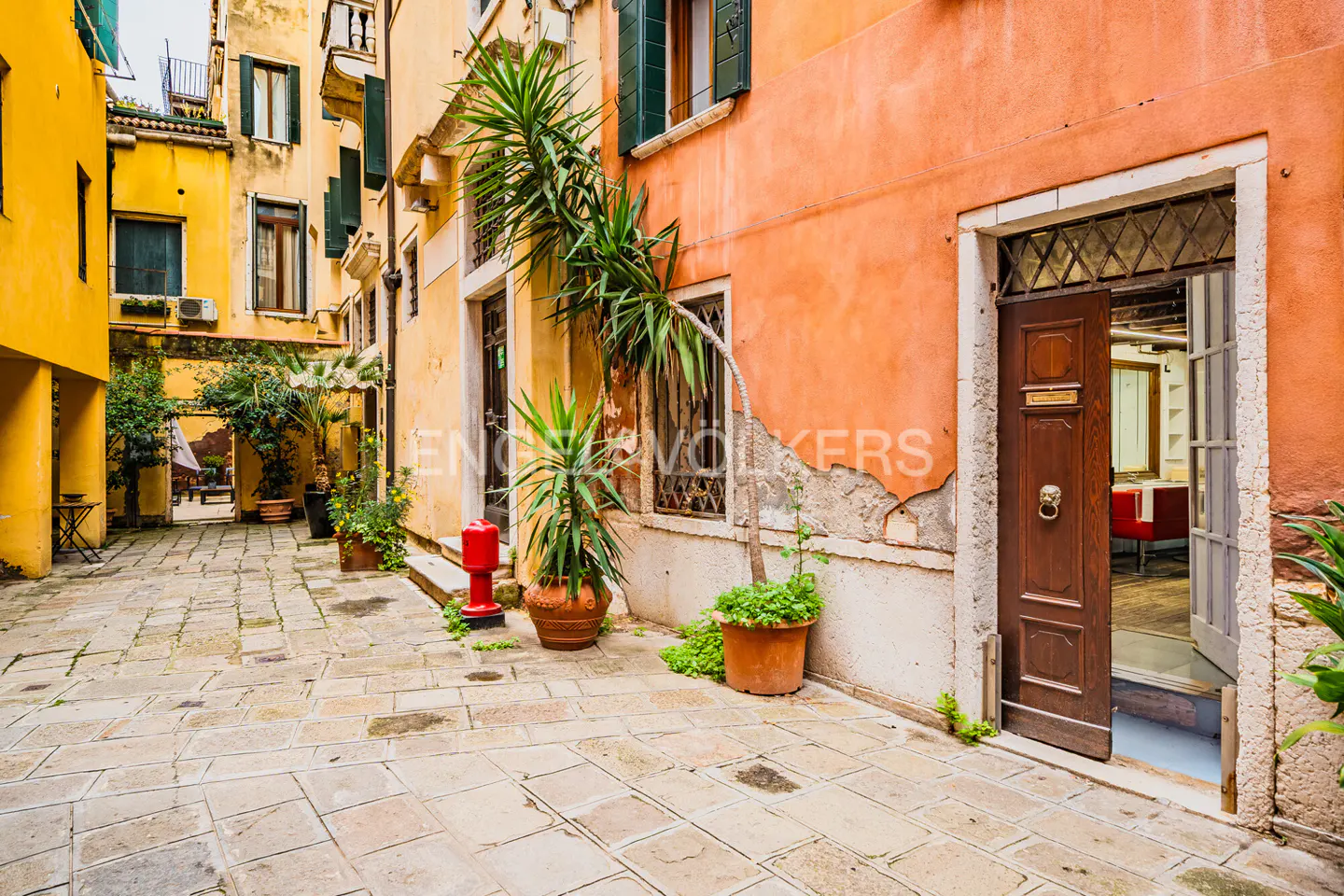 Venice street view with yellow and orange buildings, stone pavement, potted plants, and an open wooden door revealing a glimpse of a red chair inside.