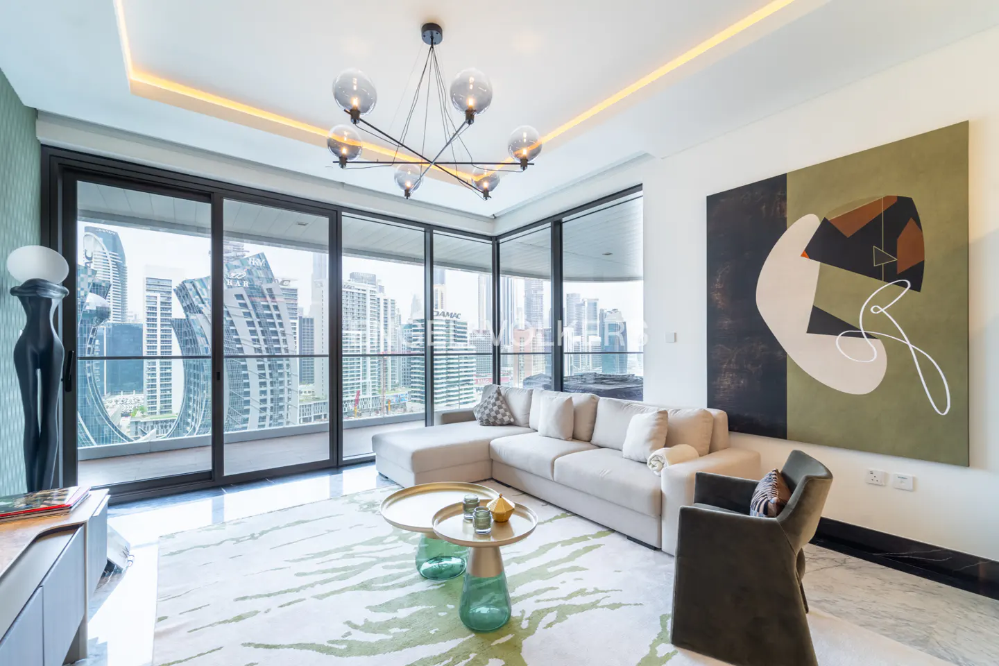 Modern living room with a beige sectional, green glass coffee tables, abstract wall art, and floor-to-ceiling city views.