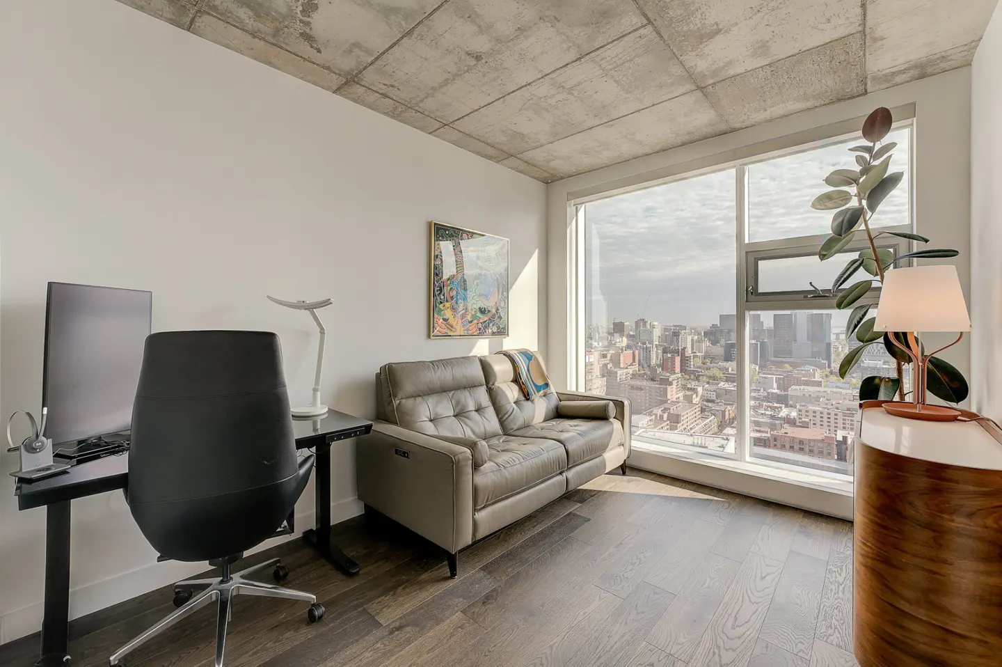 Bright, modern office space with a black desk, gray sofa, and a large window overlooking a city.