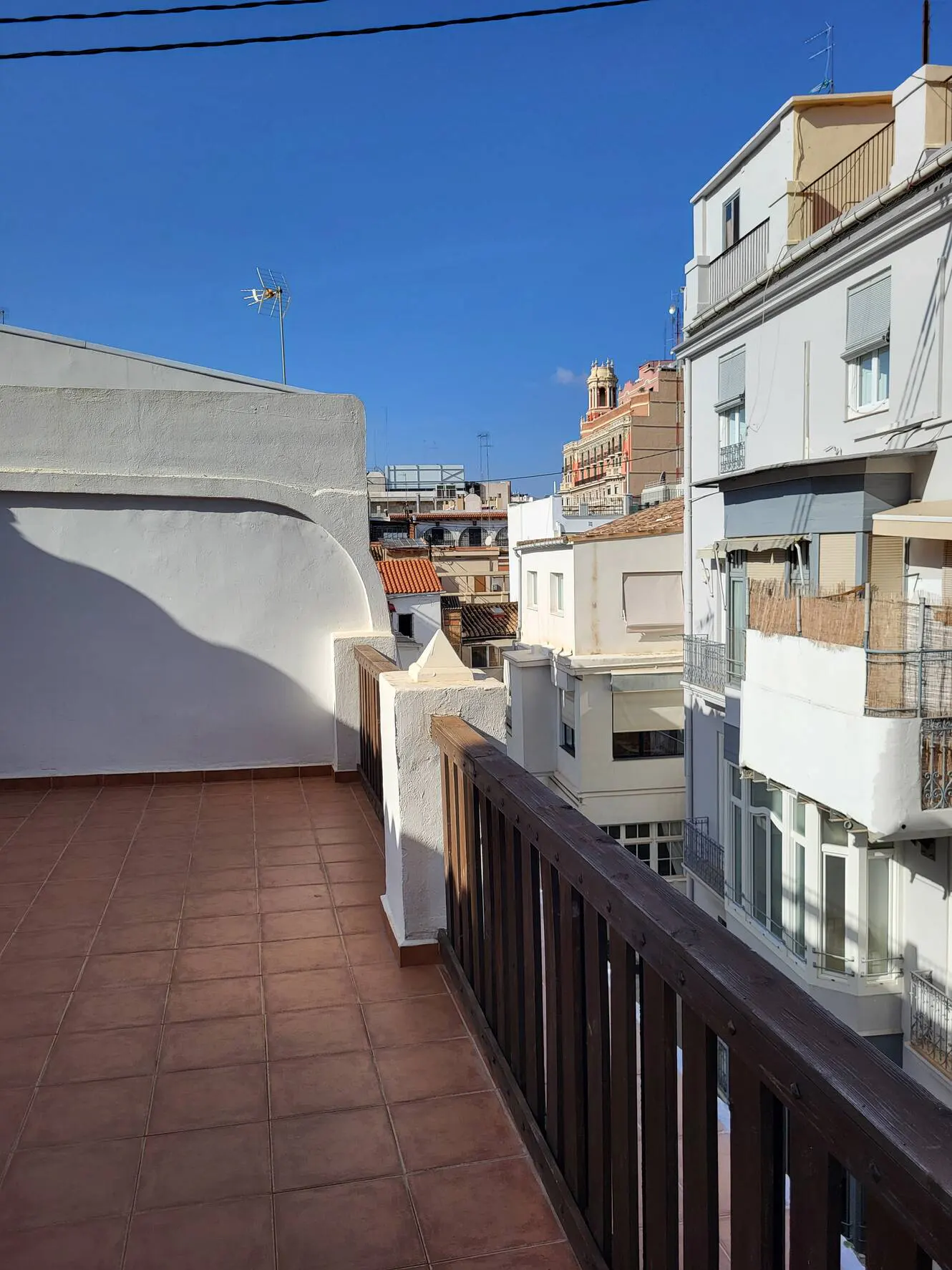 Rooftop view with brown tile floor, white walls, and dark wood railing. Buildings and blue sky in the background.