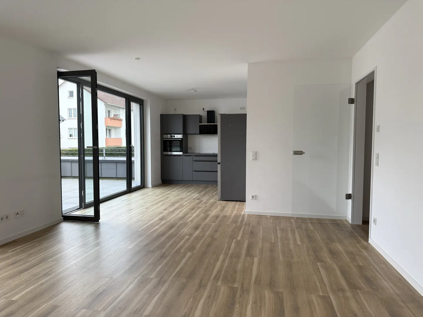 Bright, empty apartment with wood floors, white walls, and gray kitchen. Glass doors lead to a balcony.