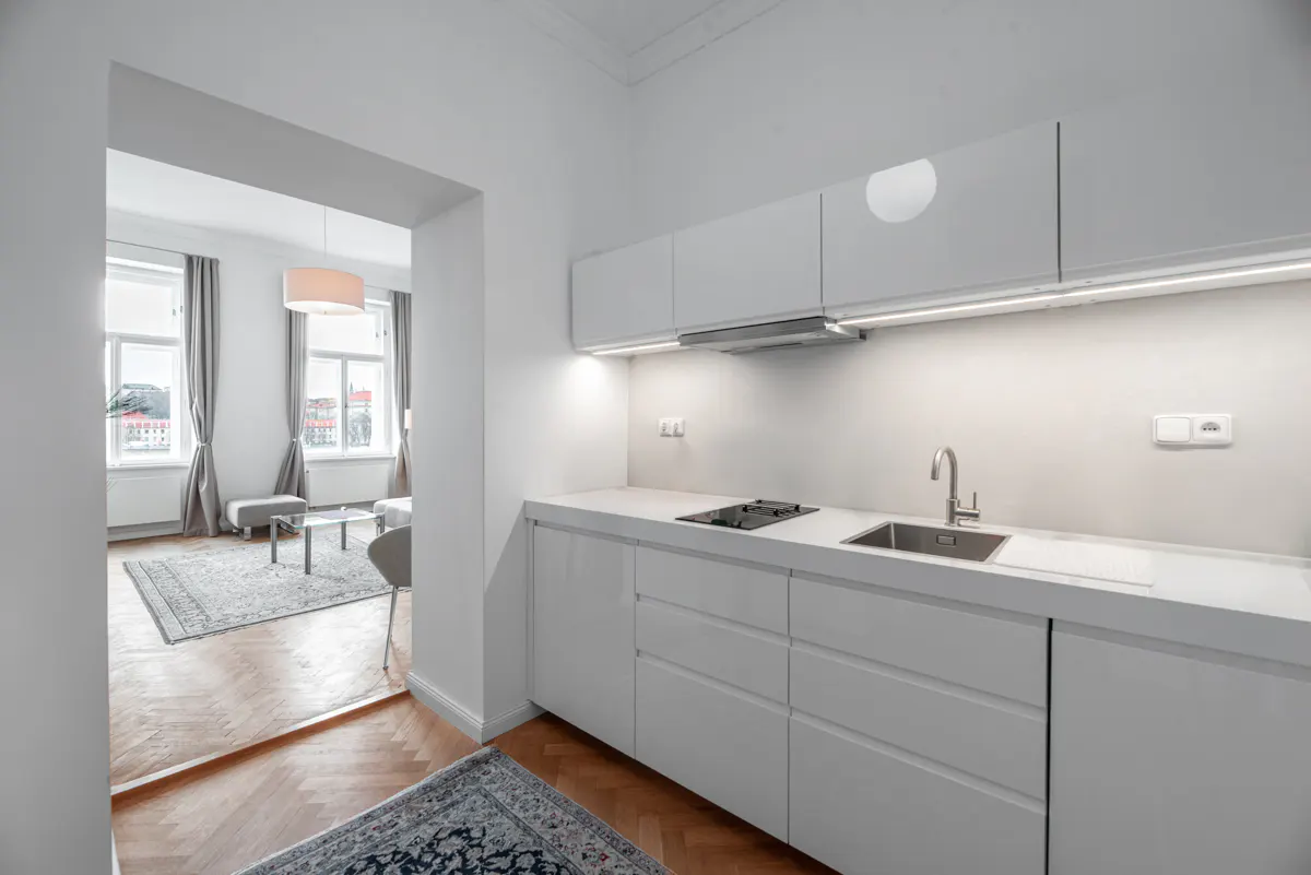 A bright, modern kitchen with white cabinets, countertops, and stainless steel sink. A living room is visible through an archway.