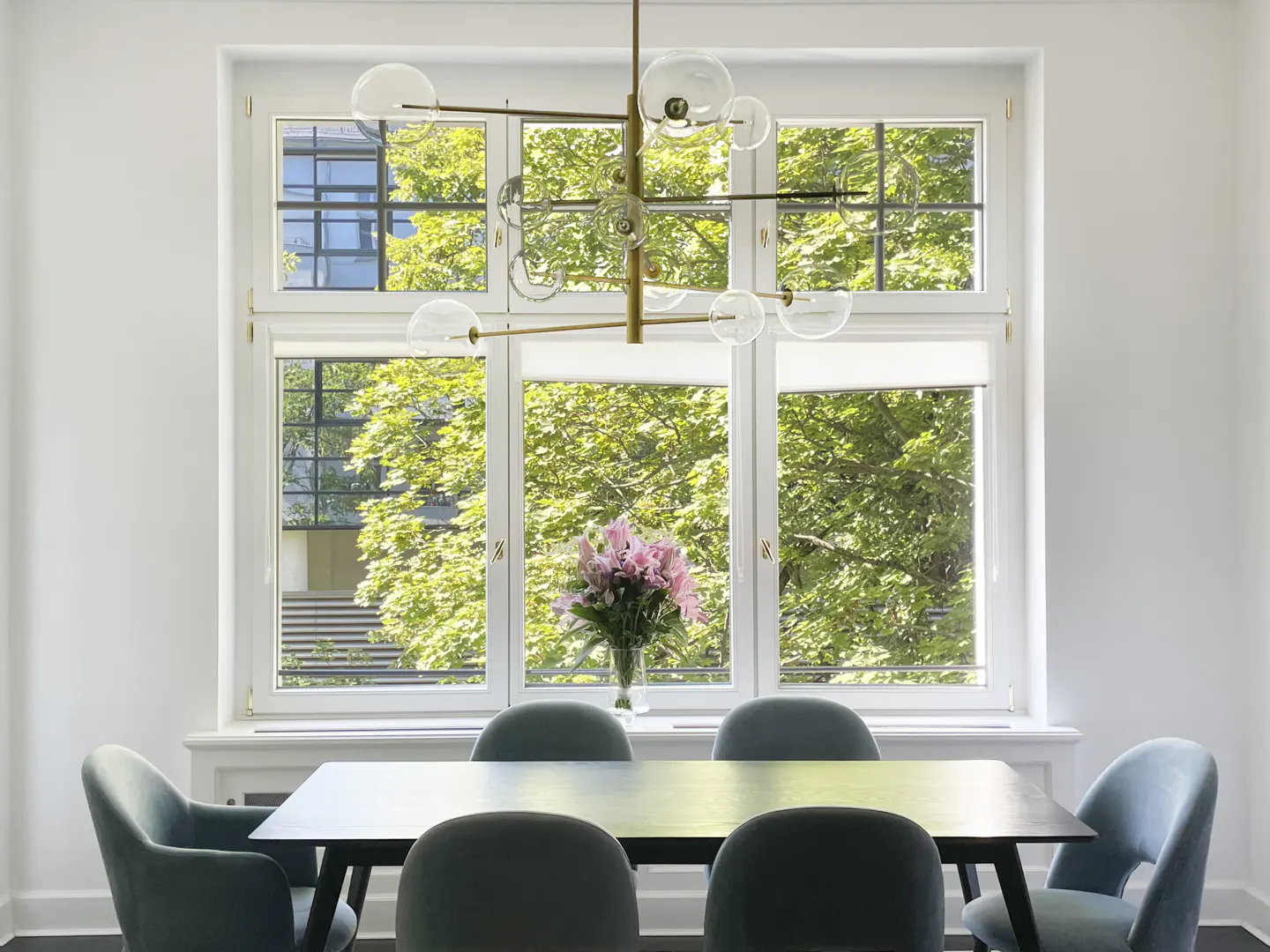 Bright dining room with a dark table, blue chairs, and a modern chandelier. A large window shows green trees outside.