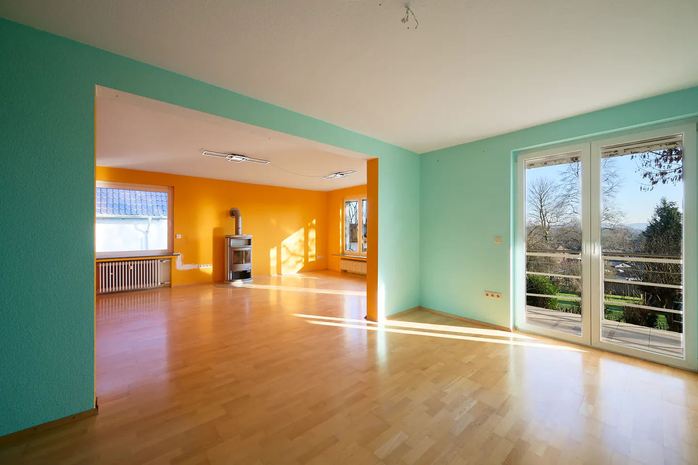 Bright, empty room with wood floors, turquoise and orange walls, and large windows overlooking a green landscape.