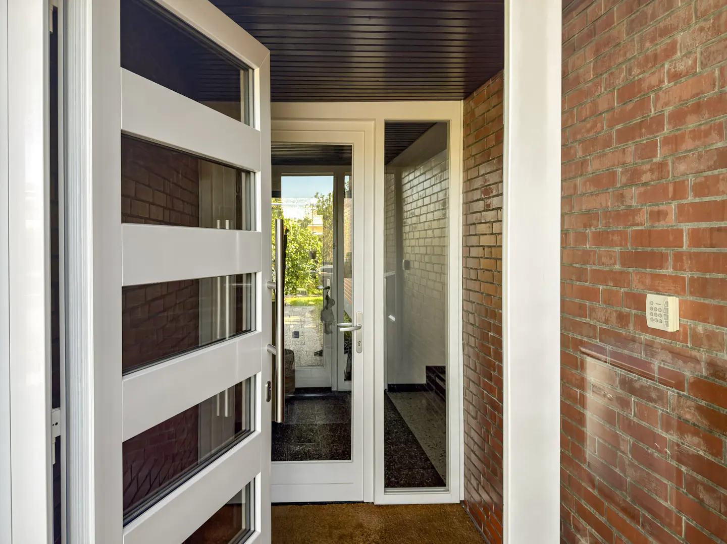 A modern white front door with horizontal glass panels stands open, revealing a bright entryway with red brick walls.