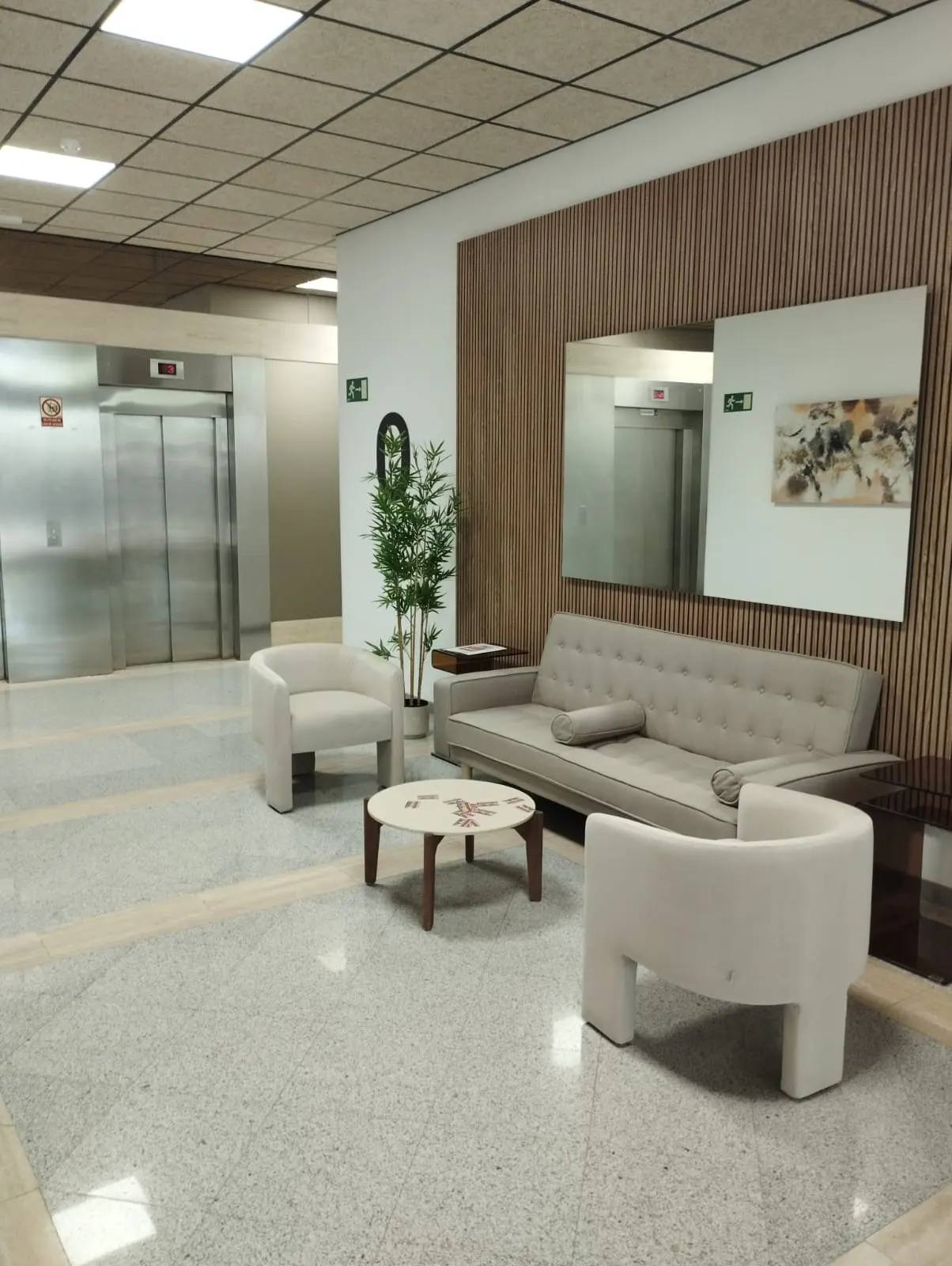 Lobby area with a gray sofa, two white chairs, and a round table. Elevators are visible in the background.