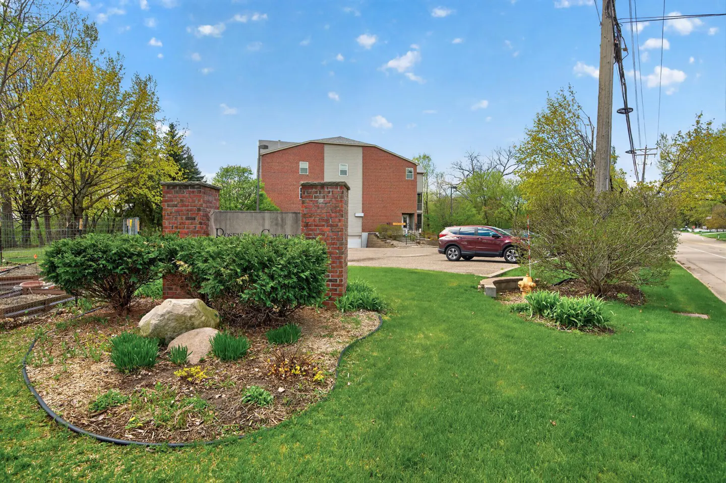 Exterior view of Rosedale Commons with brick pillars, green bushes, and a red car parked near a brick building.