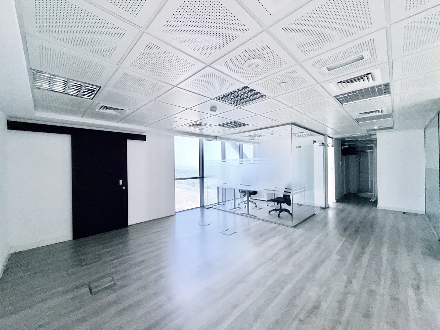 Empty office space with gray wood floors, a glass-walled conference room, and a black sliding door.