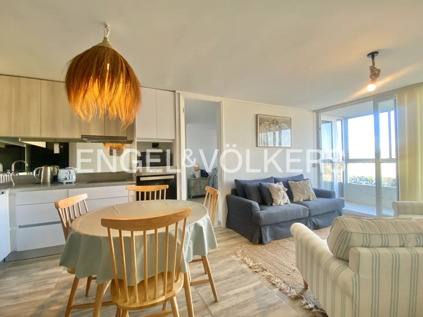 Open-concept living space with kitchen, dining table, blue sofa, and balcony with ocean view. A straw pendant lamp hangs above the table.