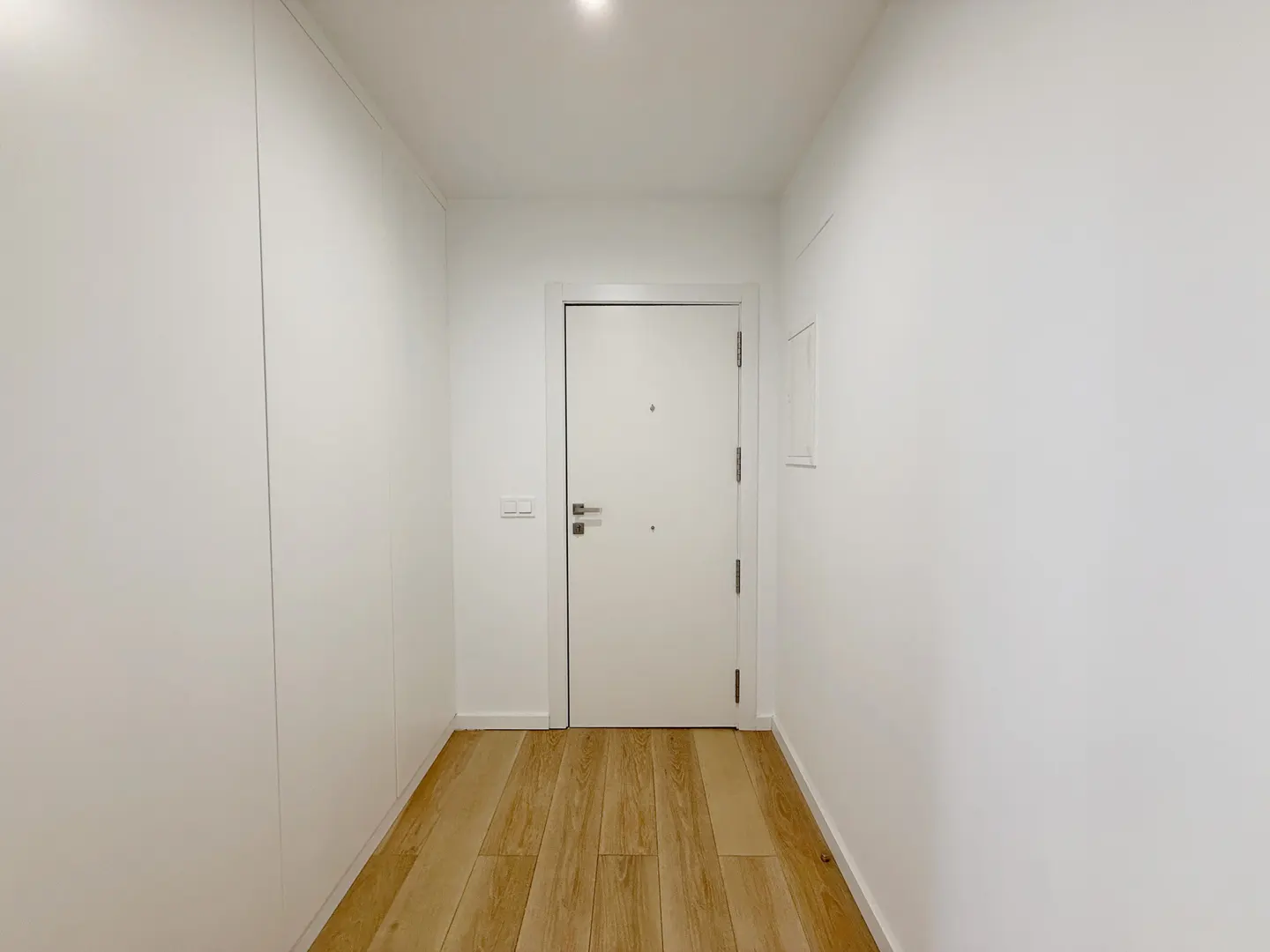 A minimalist hallway with white walls, built-in white cabinets, a white front door, and light wood flooring.