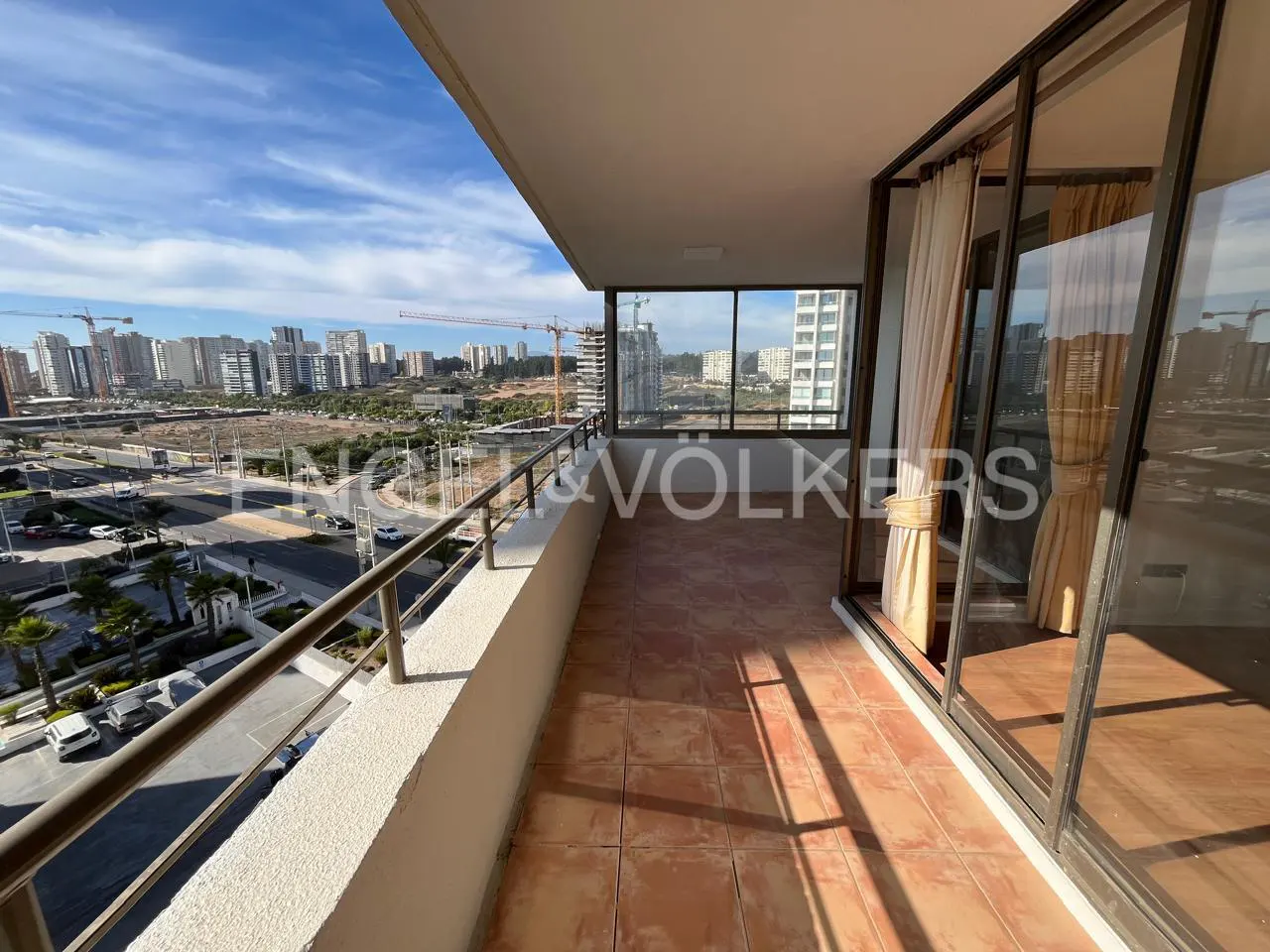 Balcony view with brown tile floor, metal railing, and city skyline. Sliding glass doors with beige curtains lead indoors. Blue sky with clouds.