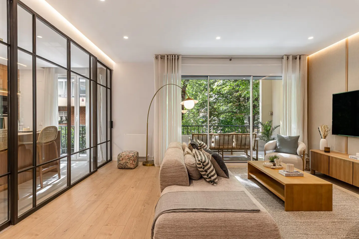 Modern living room with a beige sofa, wooden coffee table, floor lamp, and glass-walled kitchen overlooking a green balcony.
