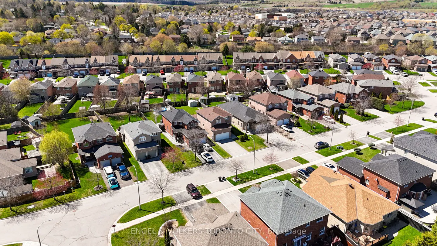 Aerial view of a suburban neighborhood with houses, green lawns, and cars parked on driveways. Trees and hills are visible in the background.