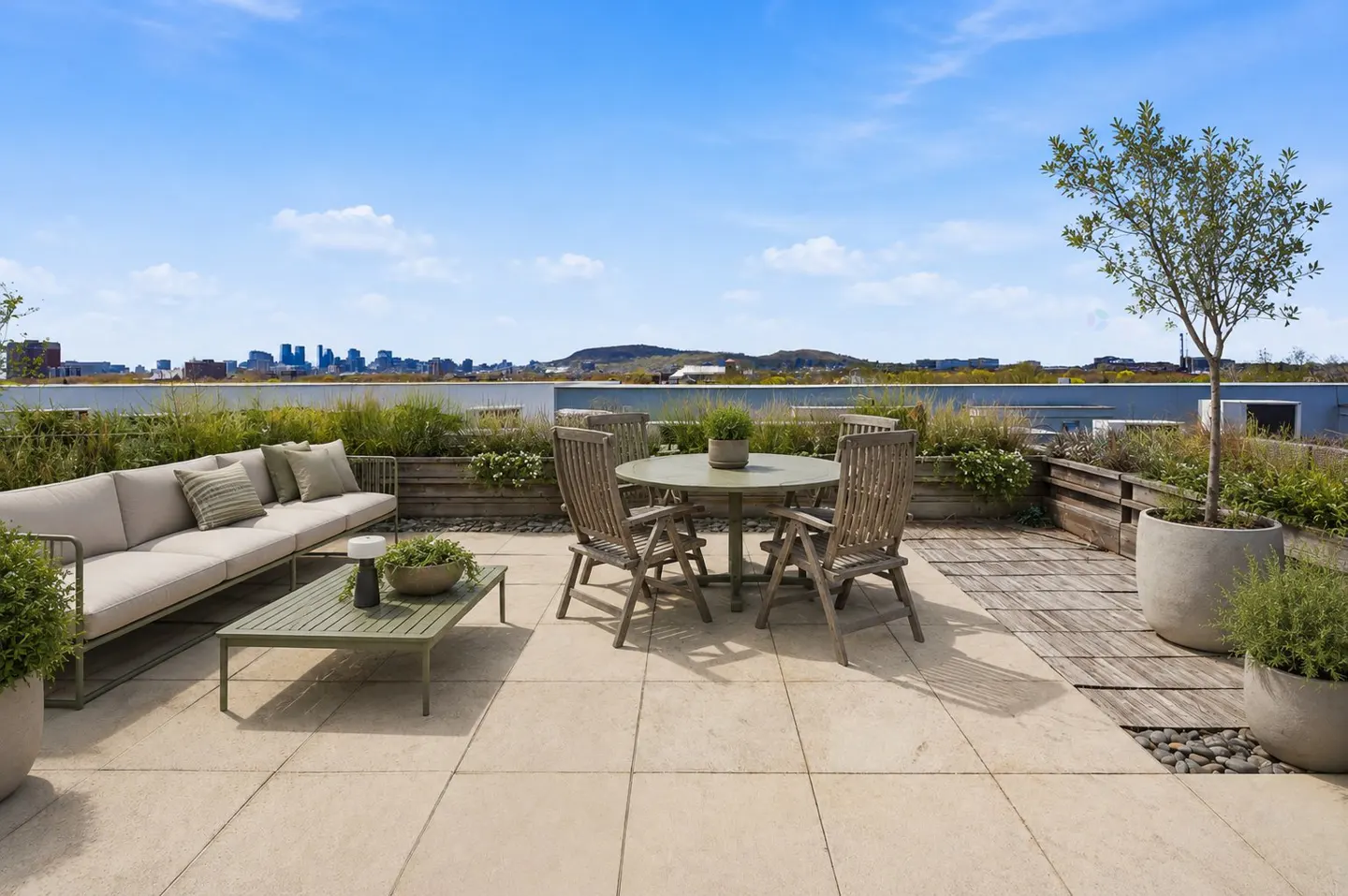 Rooftop patio with a beige sofa, green table, and wooden dining set. Planters line the edge, with a city skyline in the distance under a blue sky.