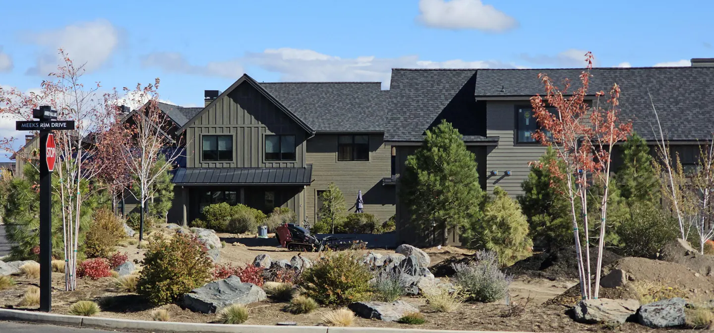 A modern, two-story house with gray siding and a dark roof, landscaped with rocks, shrubs, and small trees under a blue sky.