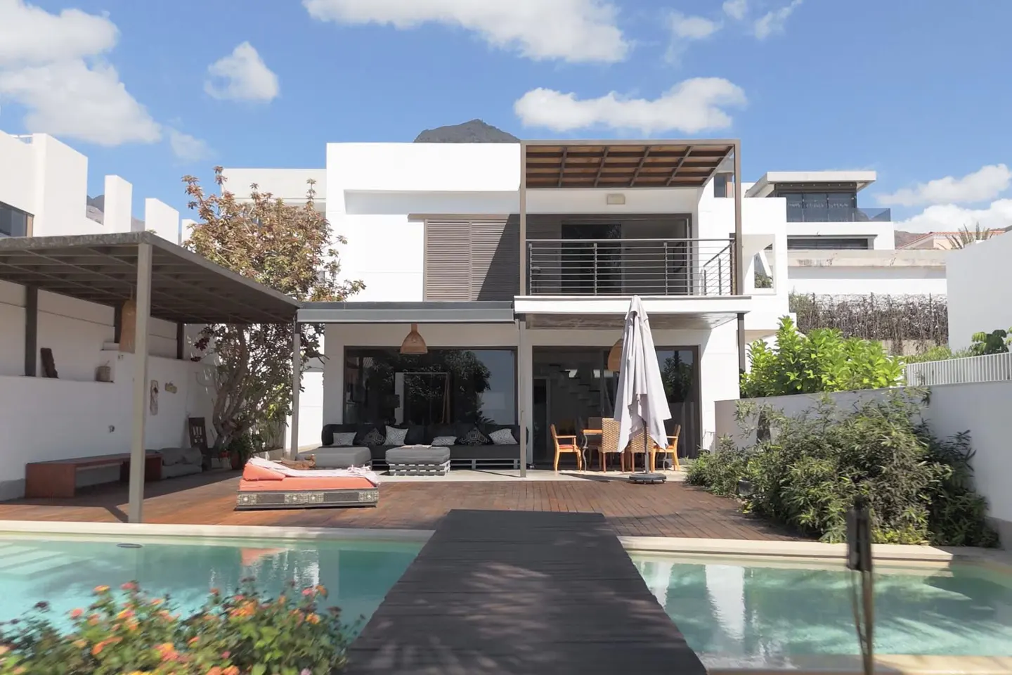 Modern white house with a pool and wooden deck. Outdoor furniture includes a lounge, dining table, and sunbed. Blue sky with clouds.