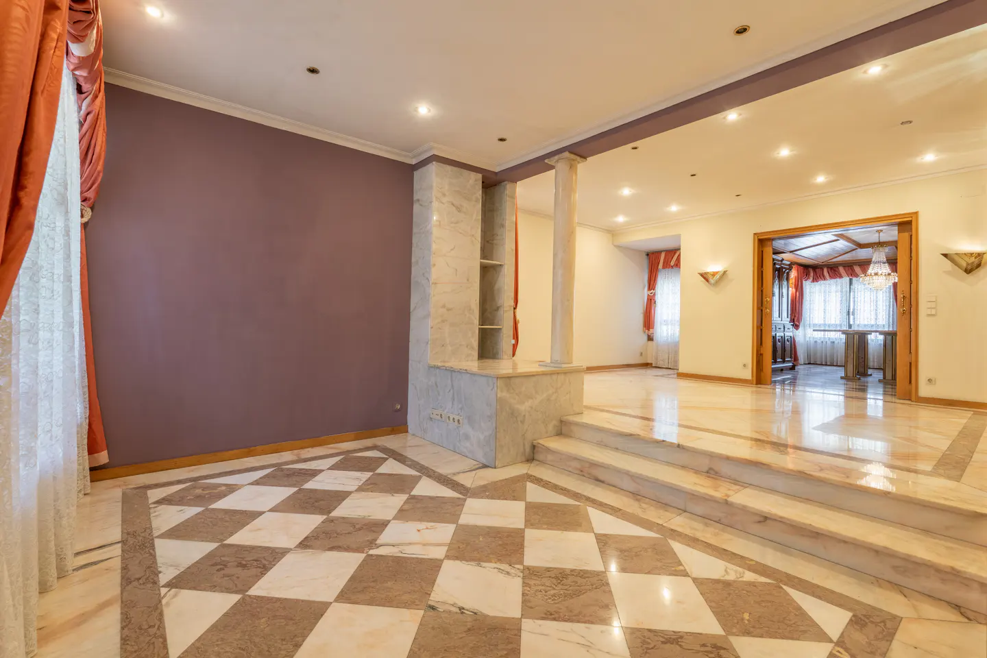 A spacious room with marble floors, a purple wall, and orange curtains. A marble pillar and steps lead to another room.