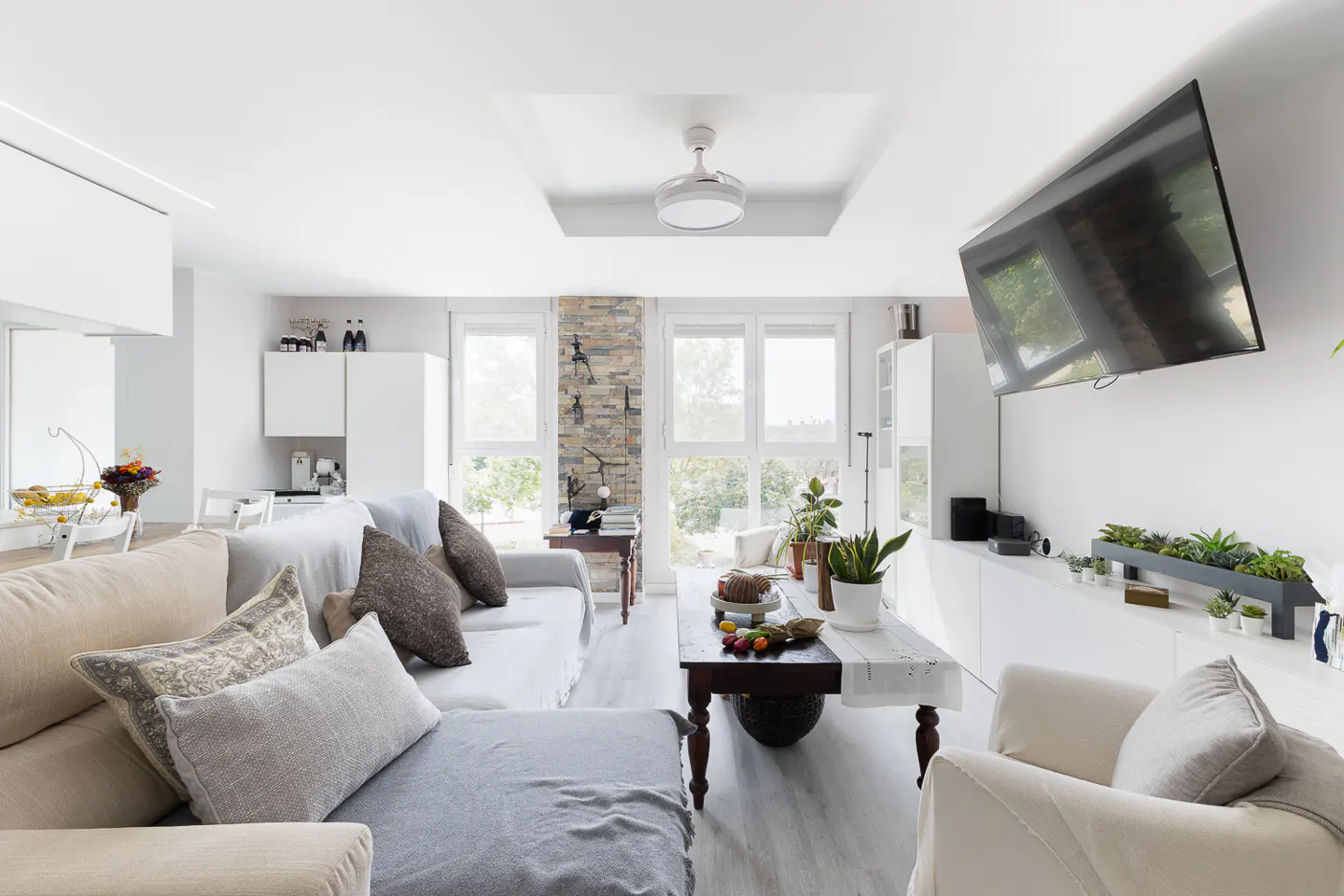 Bright living room with a beige sectional sofa, white armchair, and dark wood coffee table. A large TV hangs on the white wall. Windows offer a view of greenery.