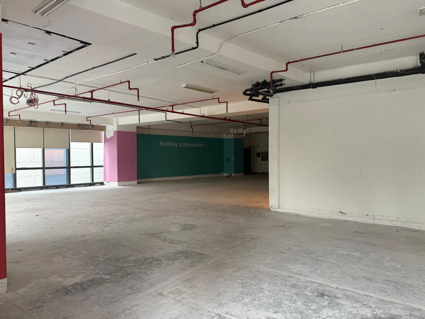 Empty commercial space with concrete floors, white walls, and exposed red and black pipes on the ceiling. A teal wall reads "Nothing is Impossible."