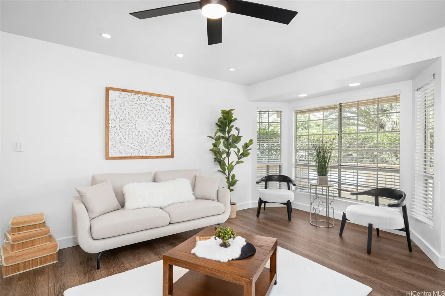 Bright living room with white walls, wood floors, and a beige sofa. A black ceiling fan hangs above, and natural light streams through the bay window.