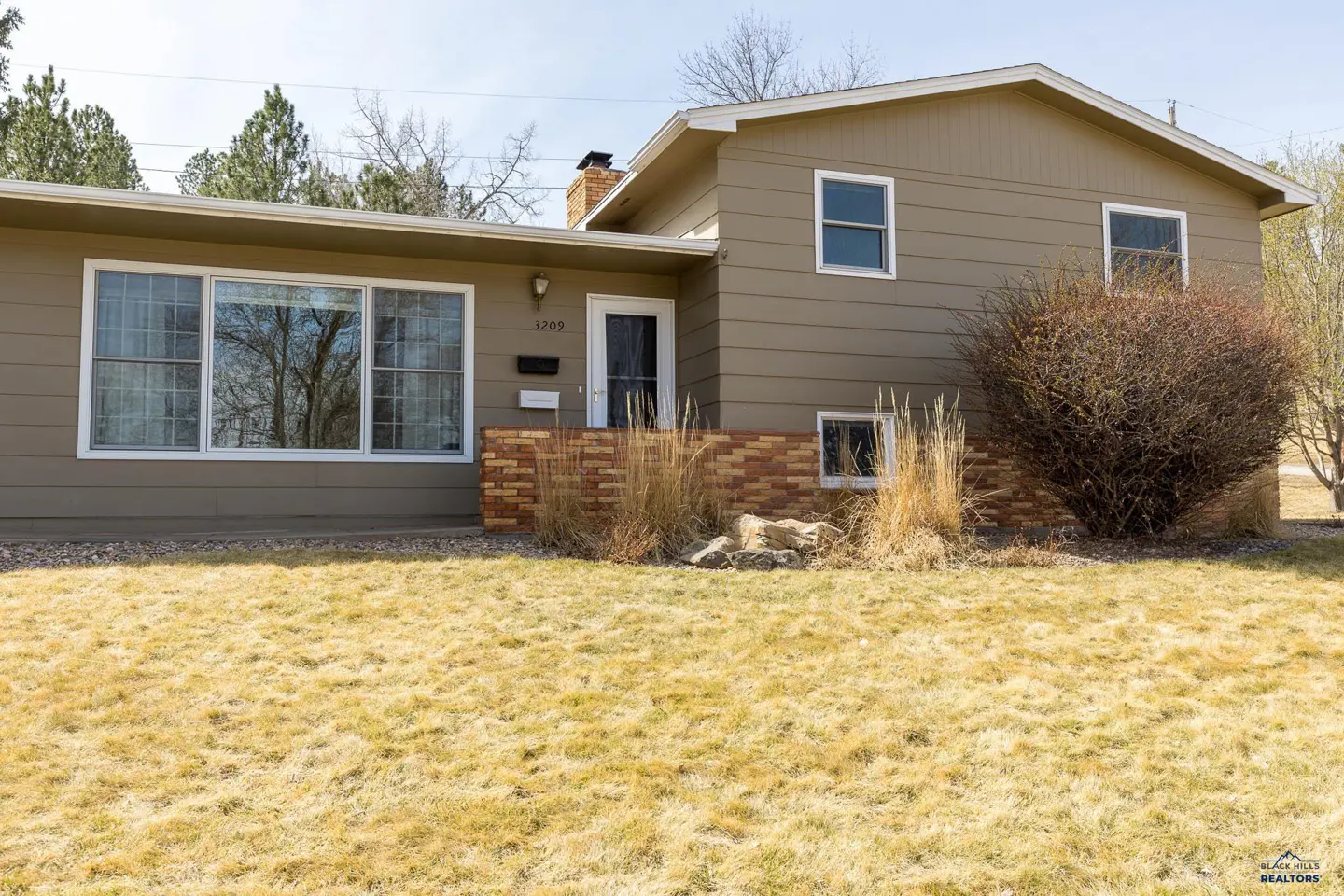 Tan two-story house with a brick foundation and a chimney. The lawn is brown and dry. There are trees in the background.