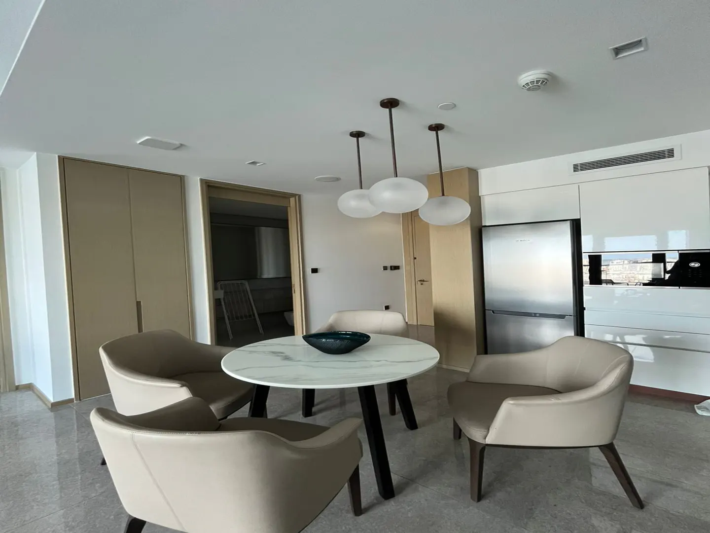 Modern dining area with a round marble table, four beige armchairs, and three pendant lights under a white ceiling.