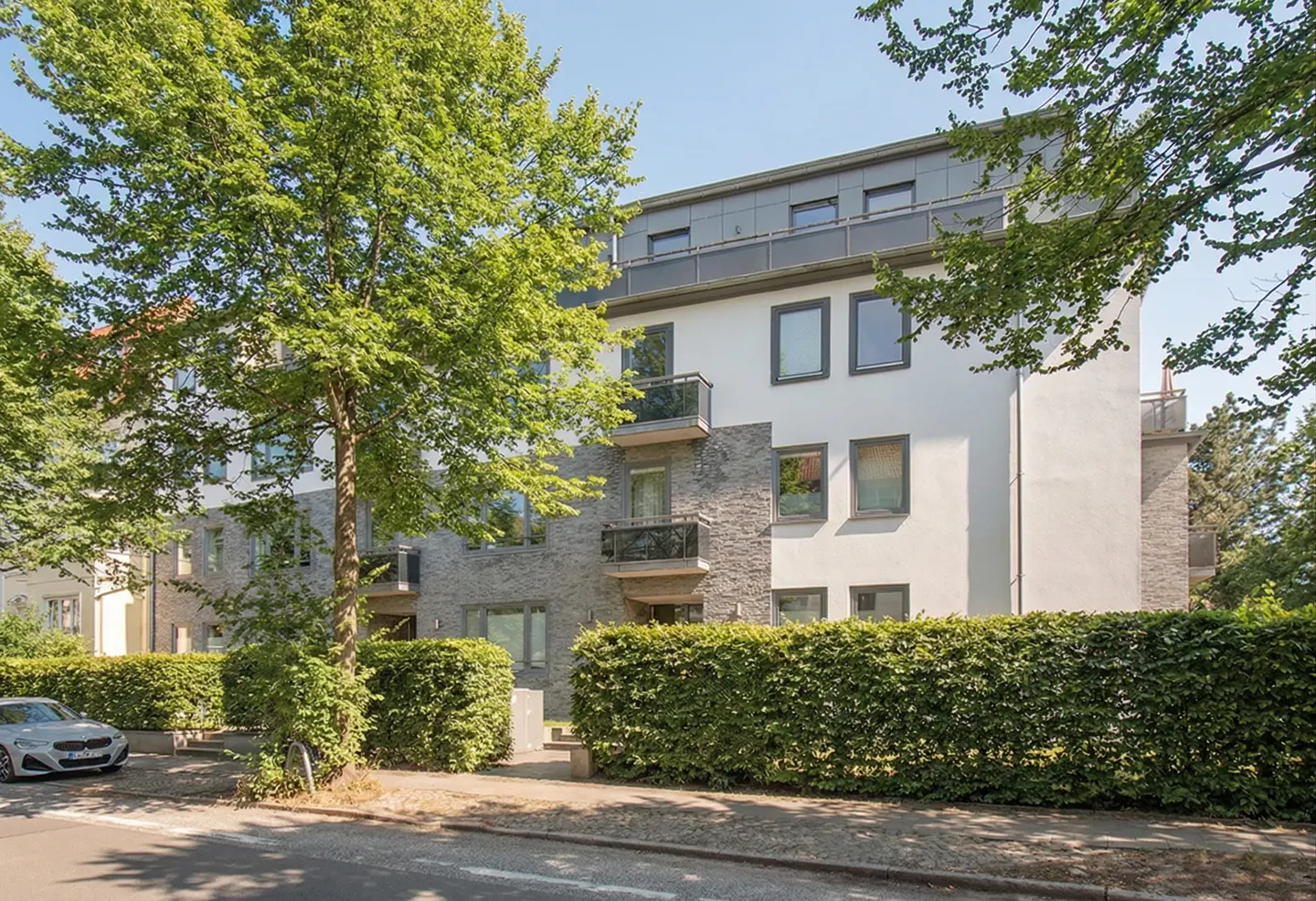 Modern apartment building with white and stone facade, balconies, and a lush green hedge in front on a sunny day.