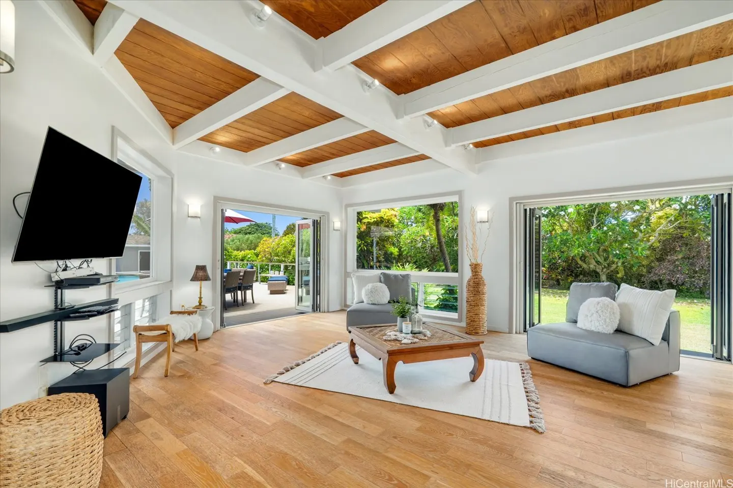 Bright living room with wood floors, white walls, and wood-paneled ceiling. Large windows and doors open to a patio and lush greenery.