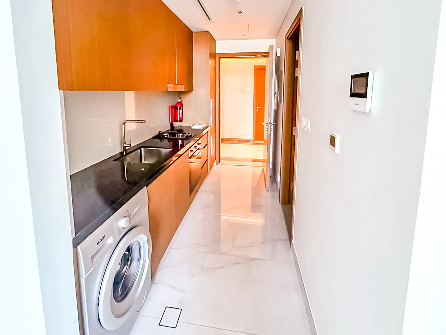 Modern galley kitchen with wood cabinets, black countertops, built-in washer, and white marble-style tile flooring.