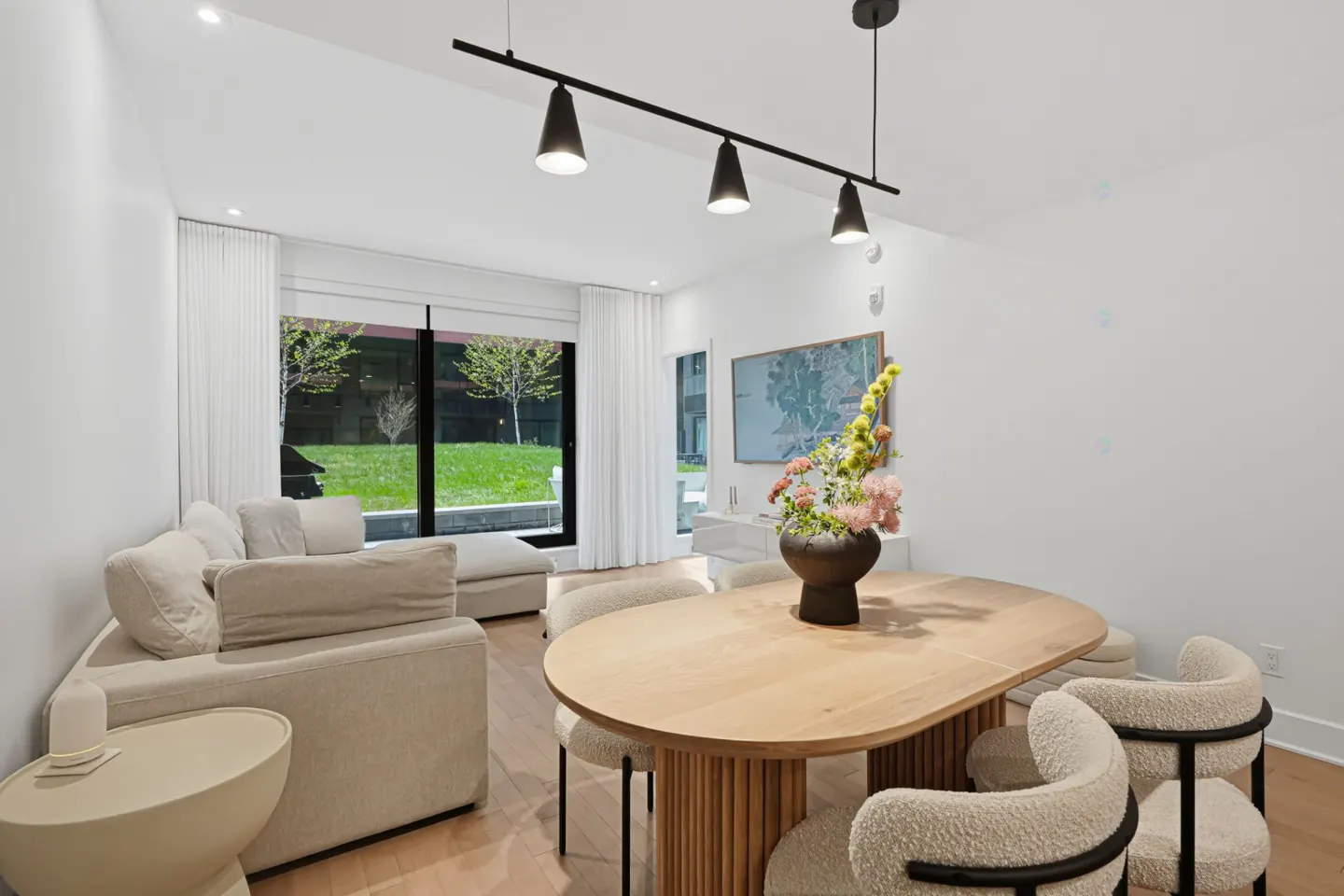 Bright, modern living room with a light wood table, six chairs, and a beige sectional sofa. A black track light hangs above the table. Large window with a view of green grass.