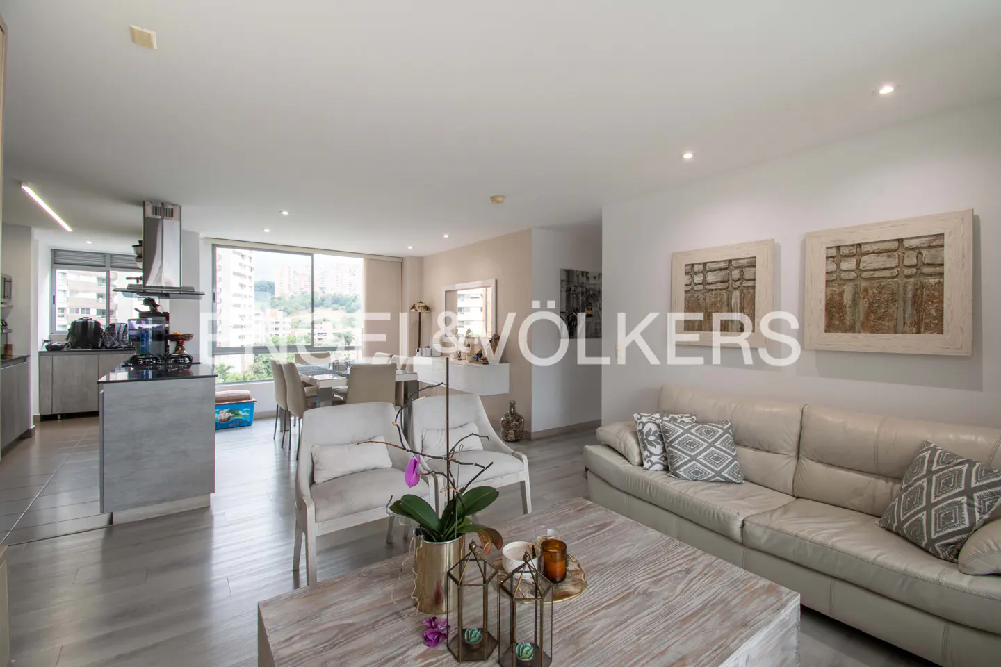 Open-concept living space with a kitchen, dining area, and living room. White sofa, chairs, and light wood floors. Artwork on the wall.