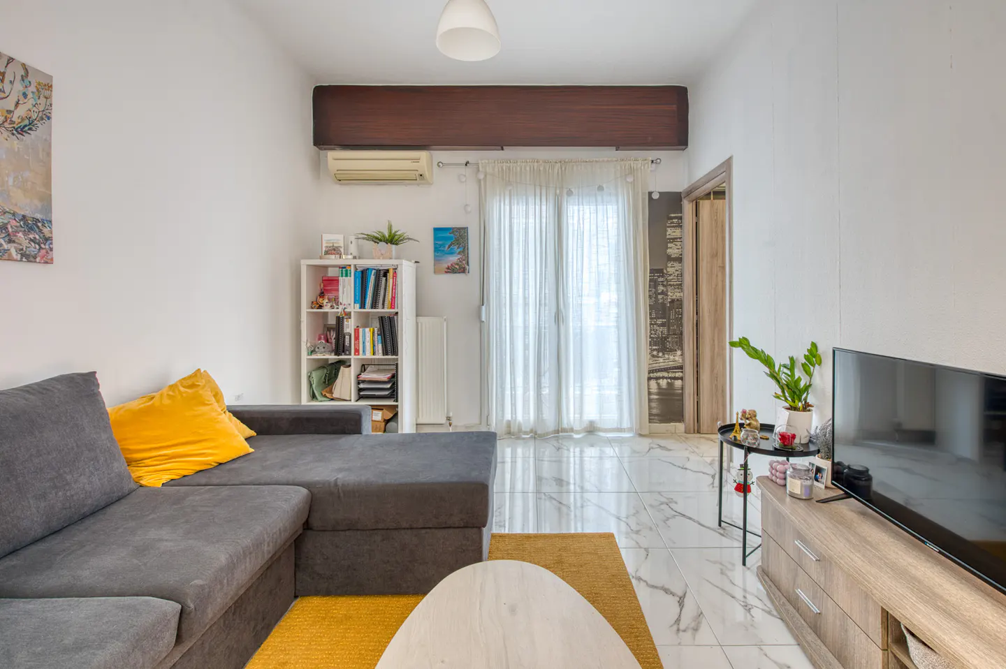 Living room with gray sectional sofa, yellow pillows, white walls, and marble floors. A white bookshelf and TV stand are also visible.