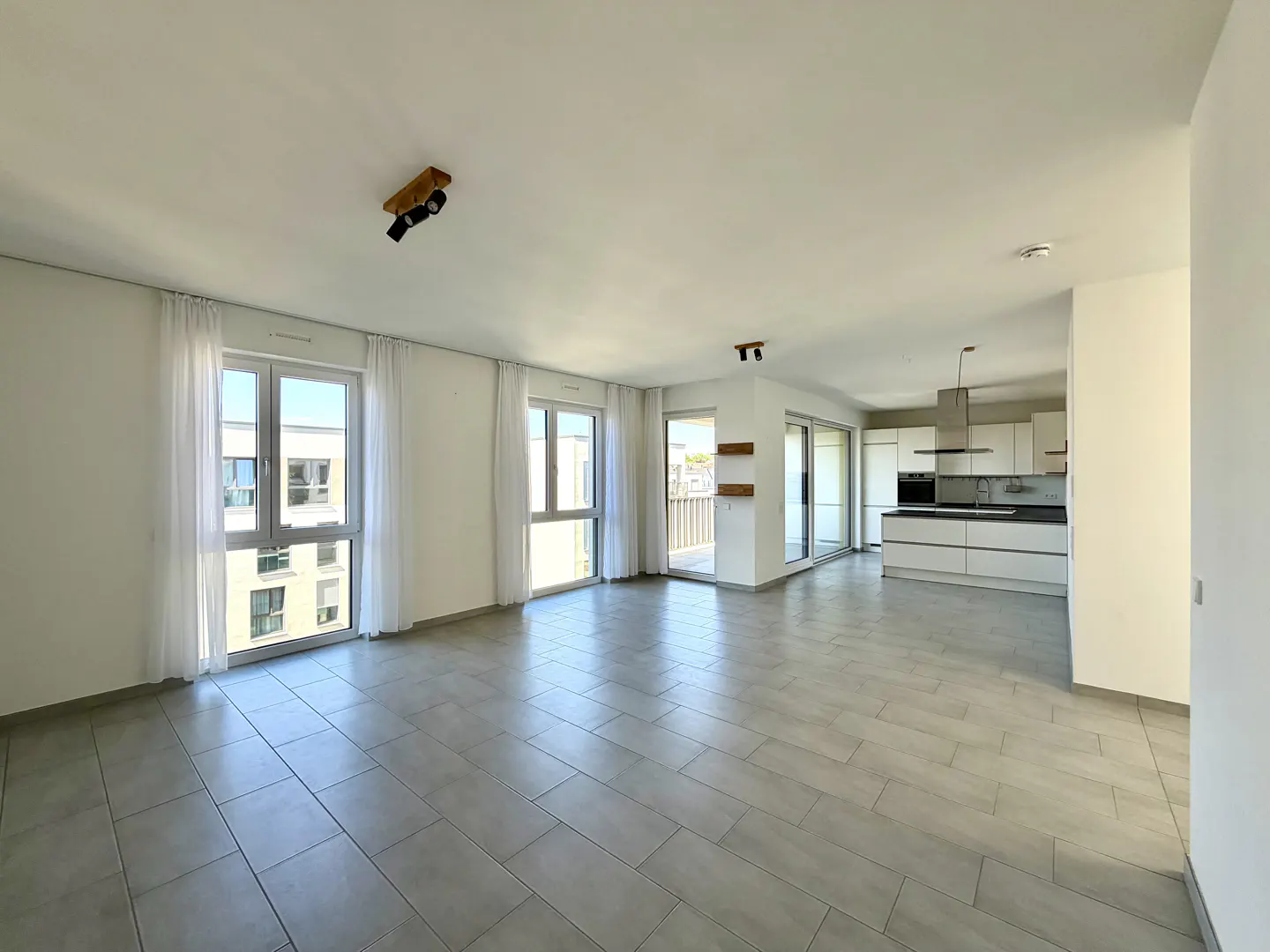 Bright, open-concept apartment with gray tile floors, white walls, and sheer curtains. A modern white kitchen is visible in the background.