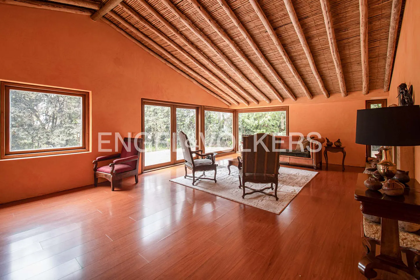 Living room with orange walls, wood floors, and a thatched ceiling. Windows overlook a green landscape. Furnished with chairs and a rug.