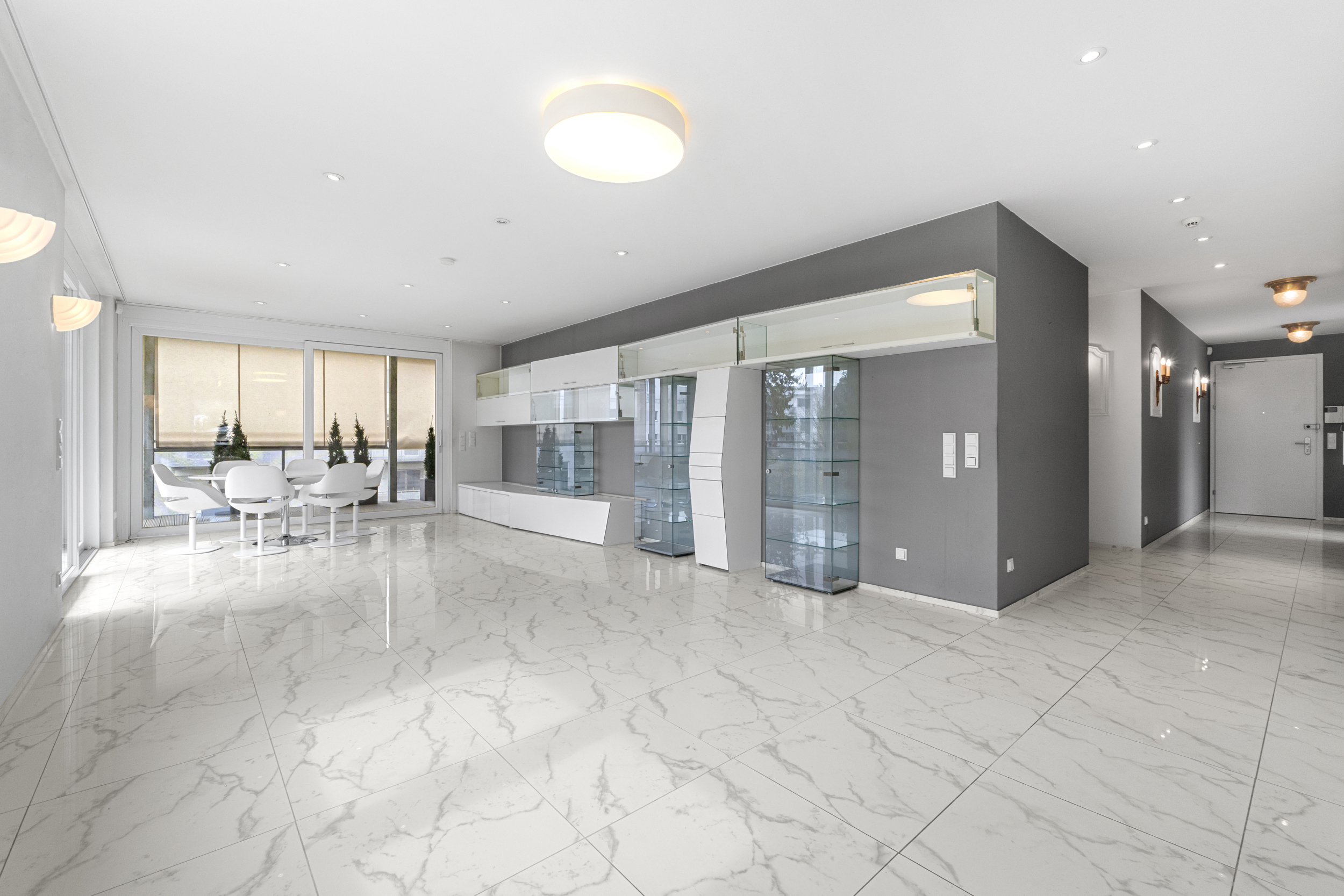 Bright, modern living room with white marble floors, gray accent wall, and glass display cabinets. Dining table near large windows.
