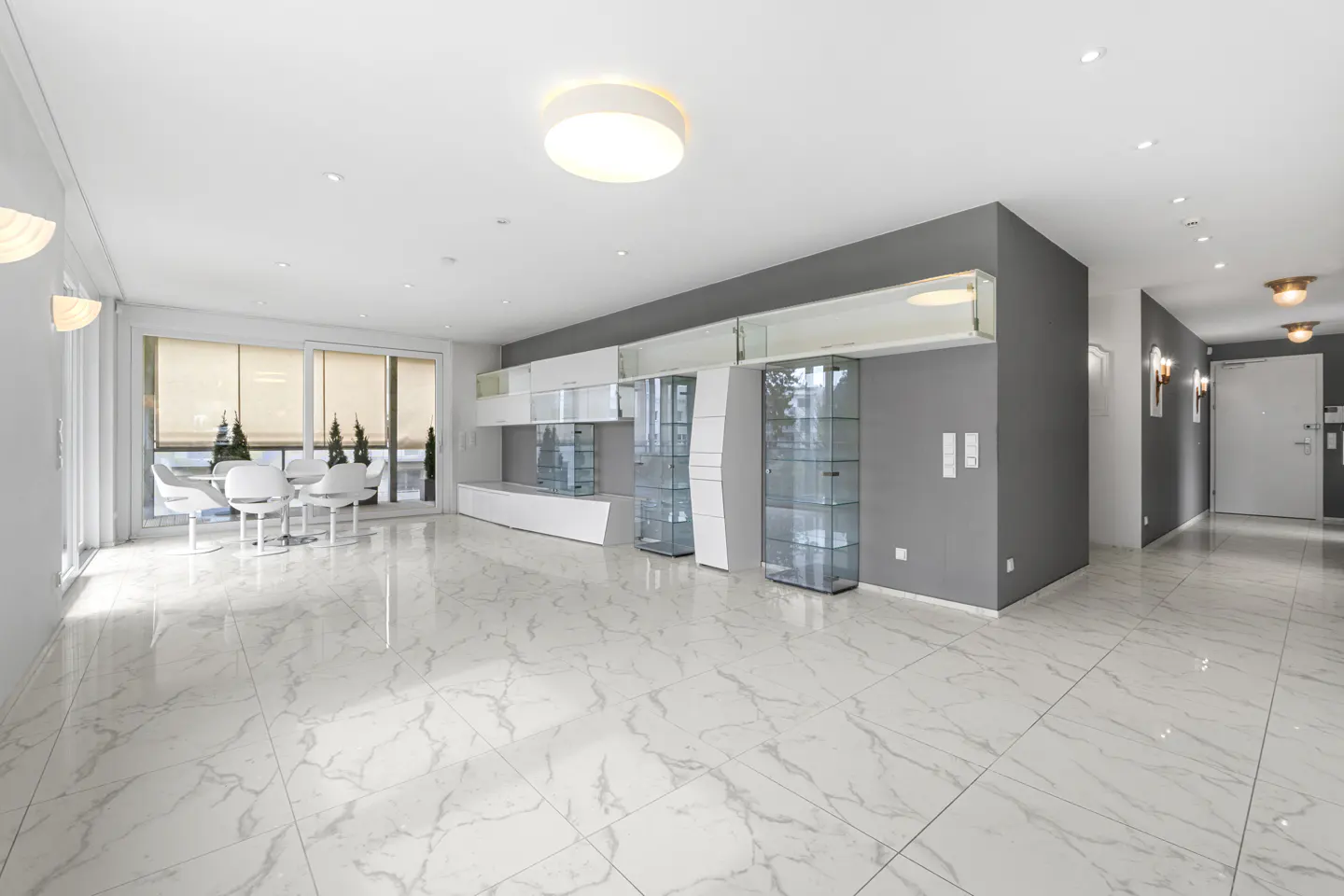 Bright, modern living room with white marble floors, gray accent wall, and glass display cabinets. Dining table near large windows.
