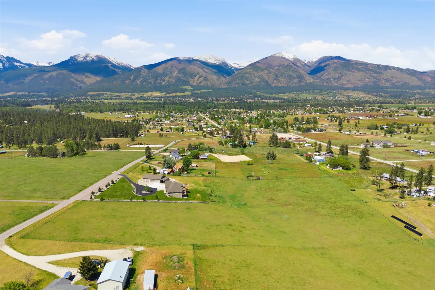 Aerial view of a green valley with houses, trees, and snow-capped mountains in the background.