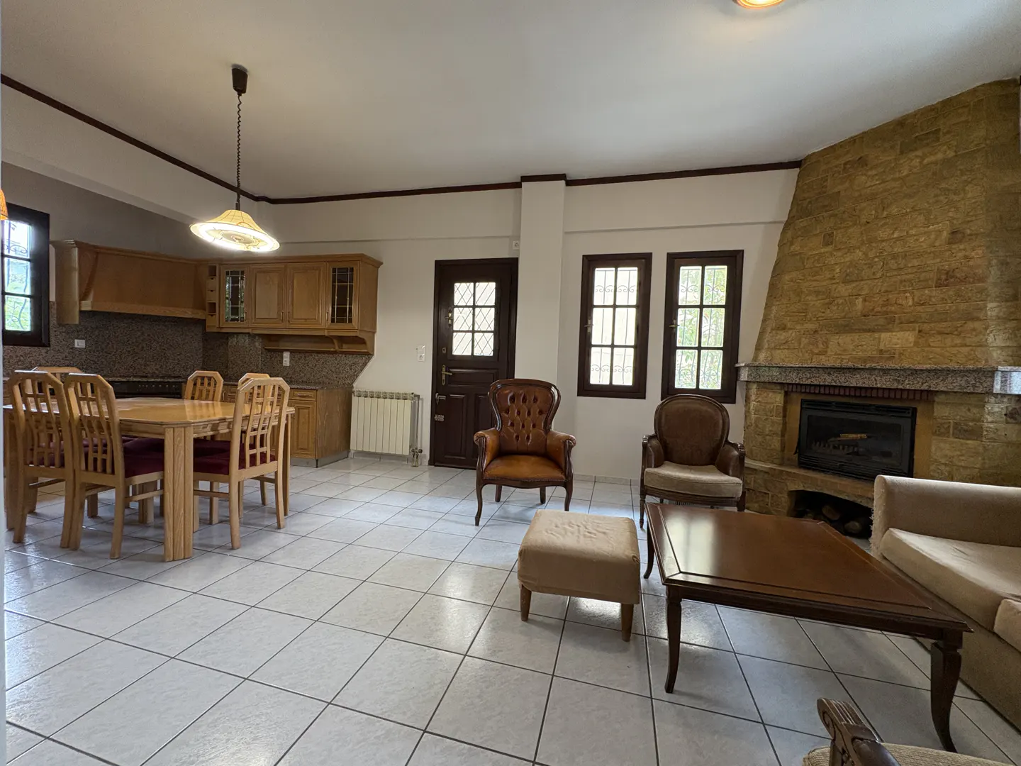 Open-concept living space with a kitchen, dining table, chairs, and a stone fireplace. Two brown armchairs and a sofa are also visible.