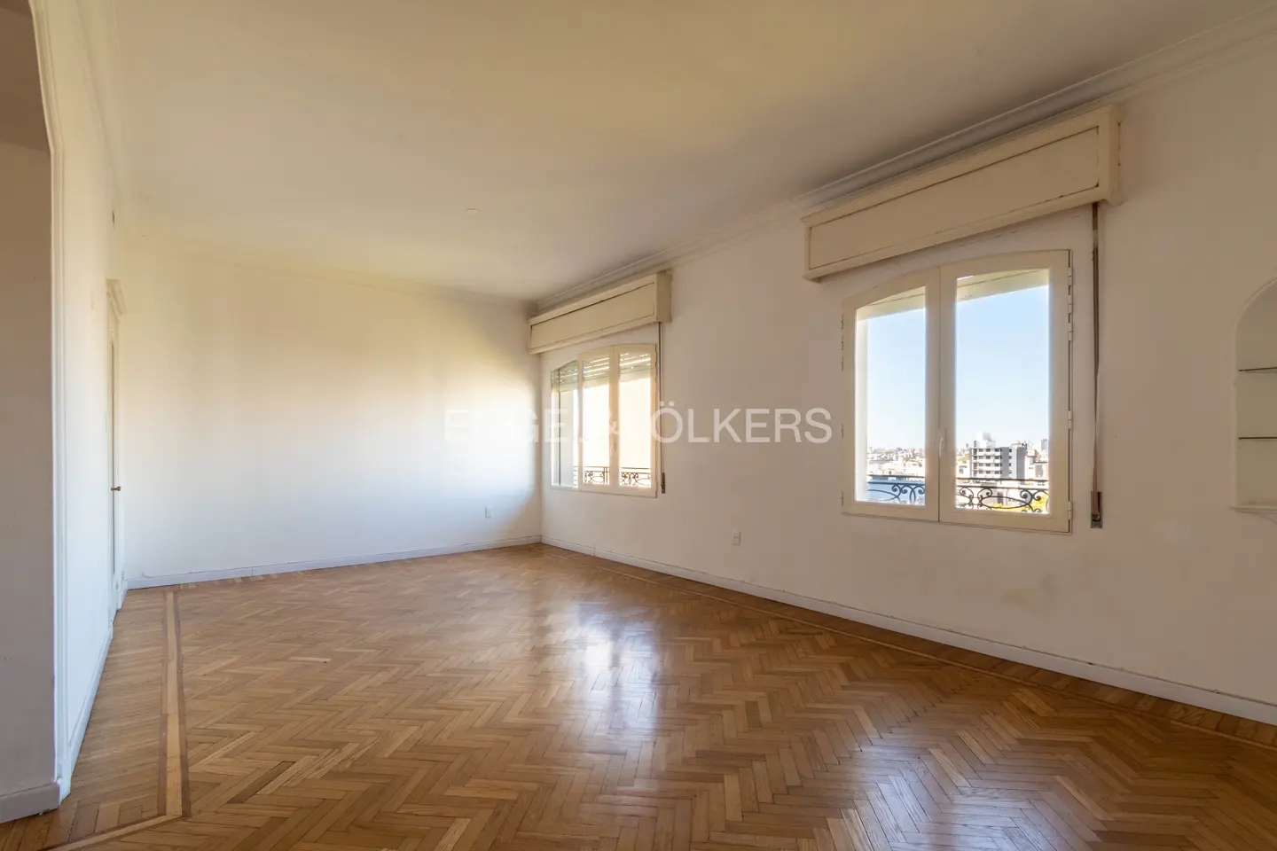 Empty room with herringbone wood floors, white walls, and two windows with city views.