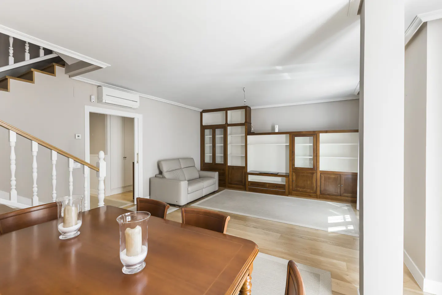 A bright living room with a wooden dining table, chairs, and candles. A gray sofa sits near a wooden cabinet. Stairs are visible in the background.