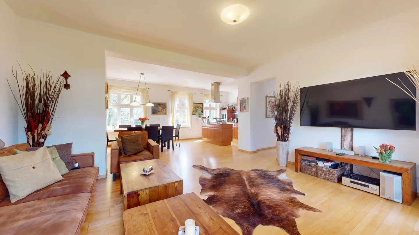 Living room with brown sofa, cowhide rug, wooden coffee tables, and a large wall-mounted TV above a wooden media console.
