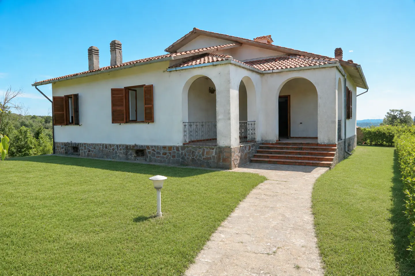 A white, one-story house with a red tile roof, brown shutters, and a stone foundation sits on a green lawn under a blue sky.