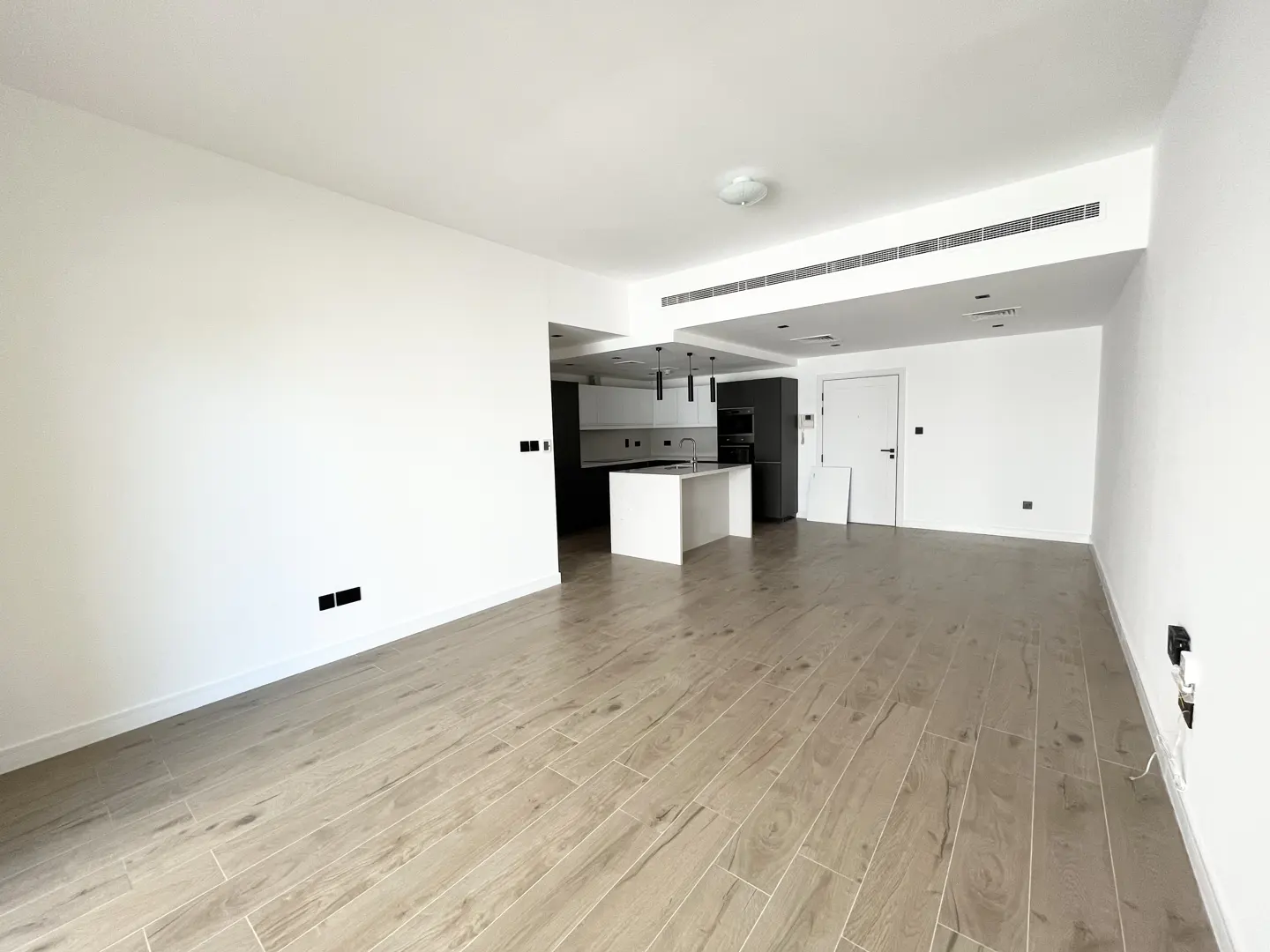 Bright, empty apartment with wood-look tile floors, white walls, and an open kitchen with a white island and gray cabinets.