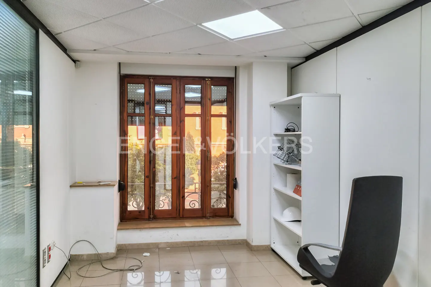 Bright office space with white walls, tile floor, and a window with brown wood frame. A white bookshelf and black office chair are visible.