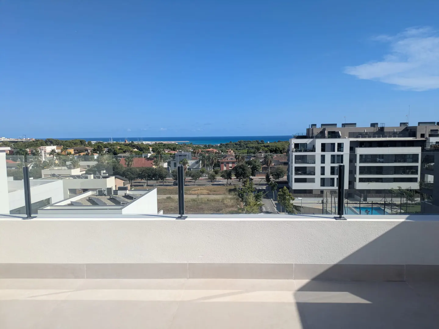 View from a balcony with a glass railing overlooking a town with buildings, trees, and the ocean under a blue sky.