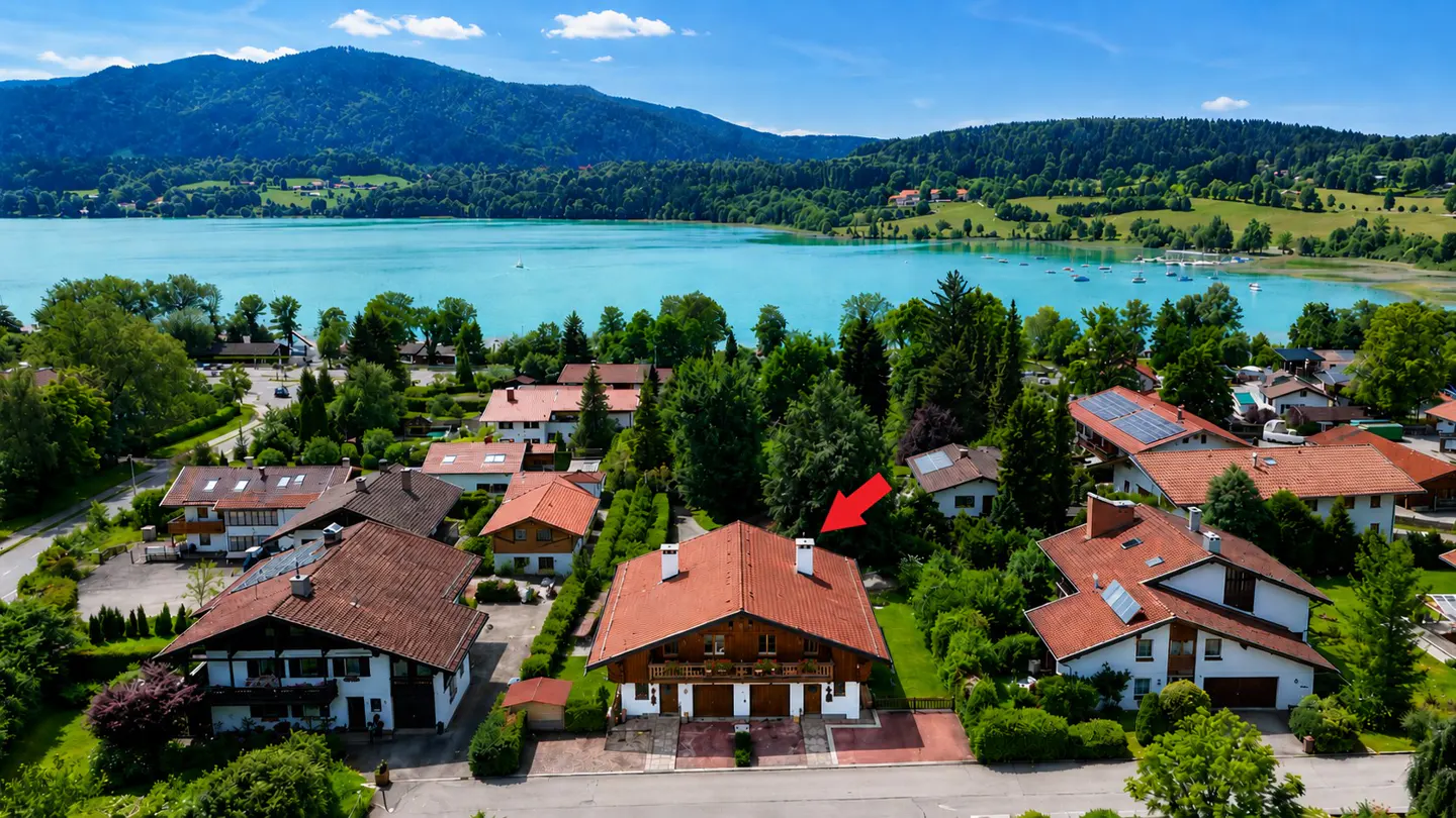 Aerial view of a house with a red roof and a red arrow pointing to it, set in a scenic lakeside town with mountains.